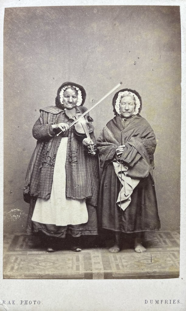 James Rae, Dumfries, ca. 1860. Zwei bettelnde Frauen stehen barfüßig im Atelier. Sie tragen beide einen Mantel und eine Haube. Die eine spielt Geige, während die andere einen Geldbeutel hält. Vielleicht ist die rechte Frau auch blind.