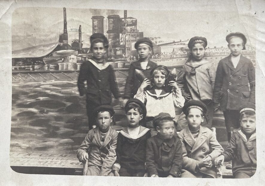 Unbekannter Fotograf, Pula, 1916. Eine Gruppe Kinder, teilweise in Marineuniform vor gemalter Kulisse. Das Kind in der Mitte hält zwei Geschosse in der Hand.