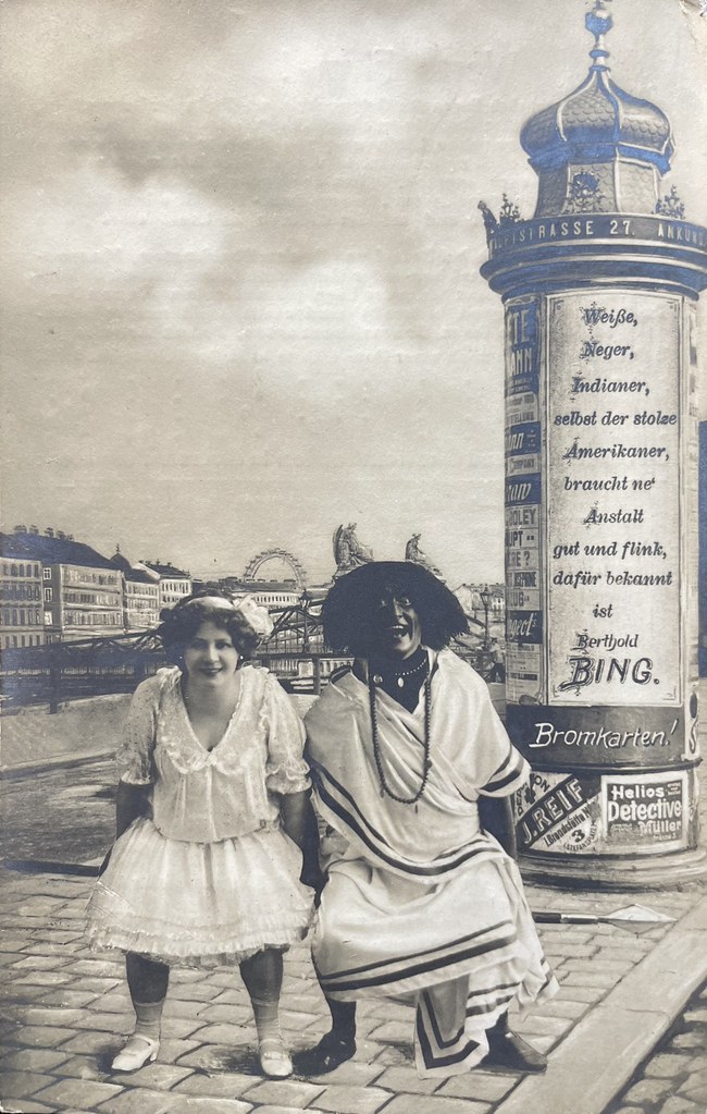 Berthold Bing, Wien, ca. 1910. Eine spannende Fotomontage, die zwei Menschen im Vordergrund und im Hintergrund die Stadt Wien zeigt. Am rechten Bildrand steht eine Werbesäule mit der Aufschrift: "Weiße, N****, Ind*****, selbst der stolze Amerikaner, braucht ne' Anstalt gut und flink, dafür bekannt ist Berthold Bing."