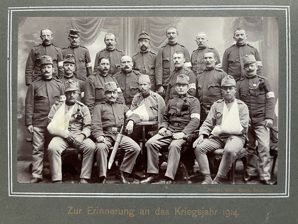 Wilhelm Mann, Salzburg, 1914. Auf dieser Fotografie sehen wir insgesamt 19 Soldaten, von denen zumindest 3 verwundet sind. Spannend ist auch, dass in der Mitte ein Soldat mit Fez sitzt, vermutlich aus einem bosnischen Regiment. Unter der Fotografie steht „Zur Erinnerung an das Kriegsjahr 1914.“
